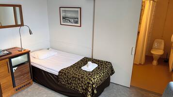 Ett hotellrum med en säng, ett skrivbord med en lampa, en liten matta och ett badrum som syns genom en öppen dörr.