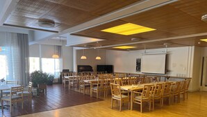 A spacious dining area with wooden tables and chairs, a piano, and a large screen on the wall.