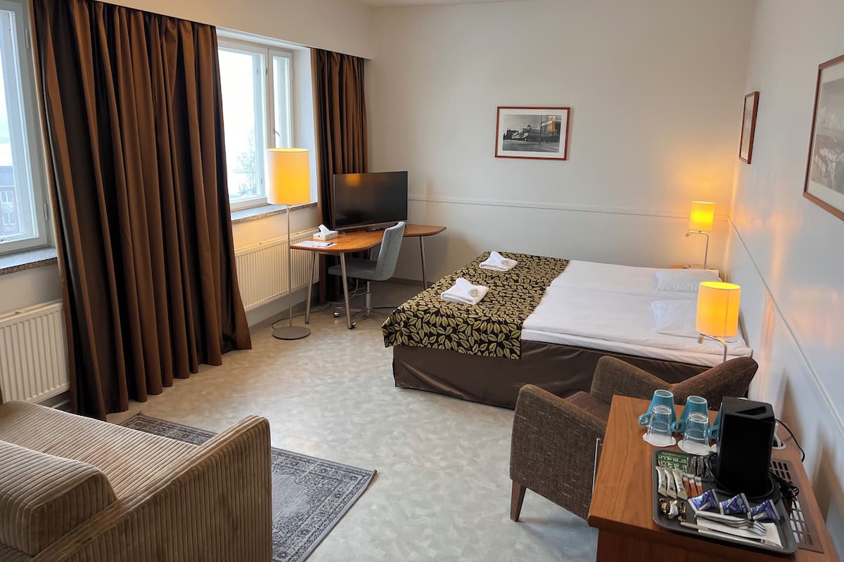 A hotel room with a bed, a desk with a computer, a TV, a chair, a sofa, and a small table with cups and snacks.