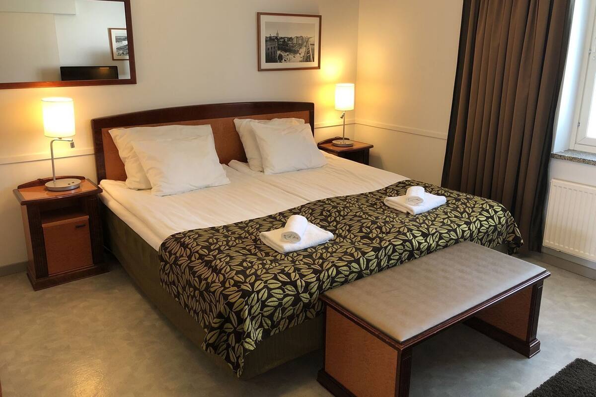 A hotel room with a bed, two bedside tables, a bench, a mirror, and a framed picture on the wall.