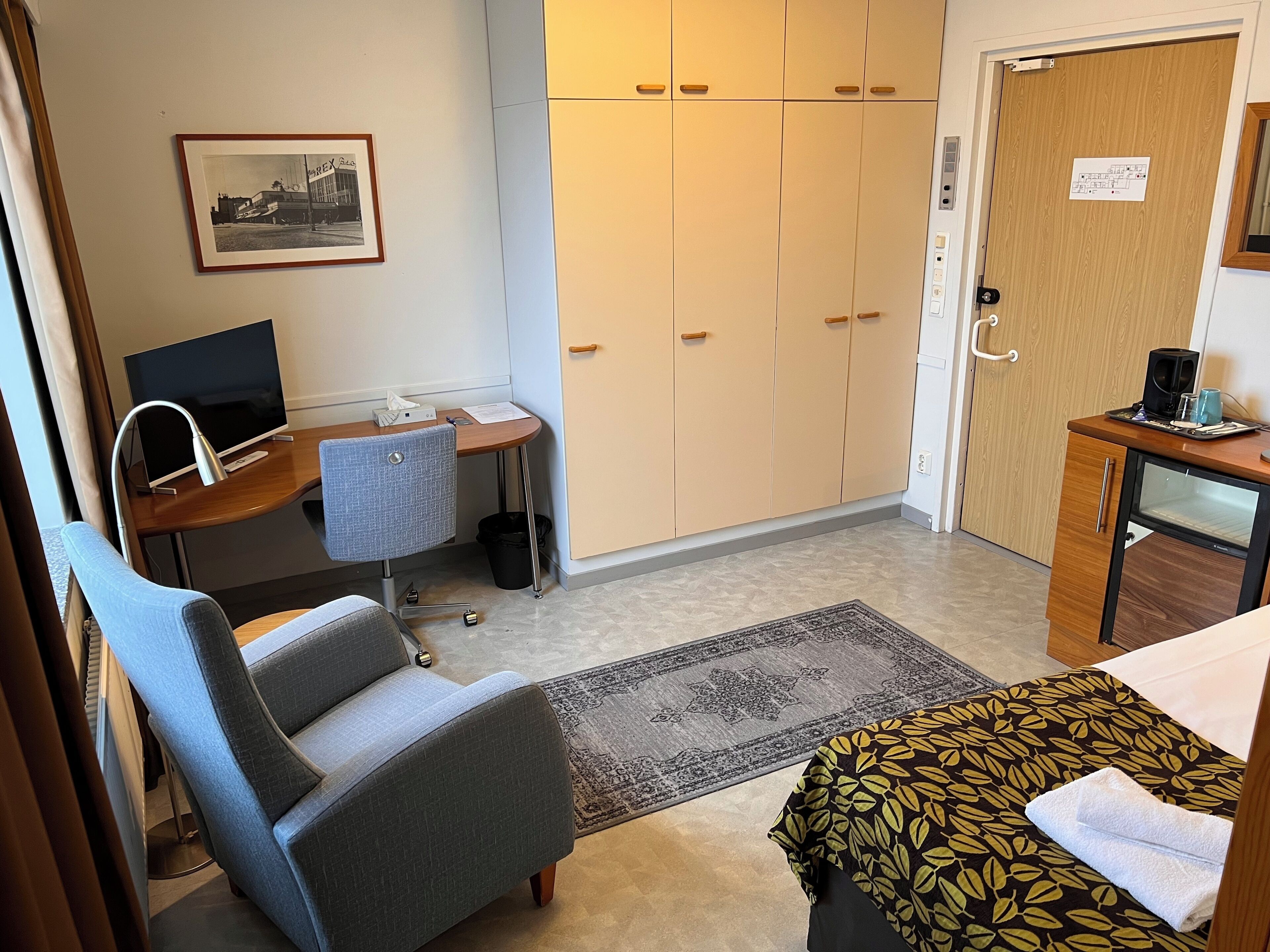 A hotel room with a bed, a desk with a computer, a chair, a wardrobe, and a small table with a coffee maker.