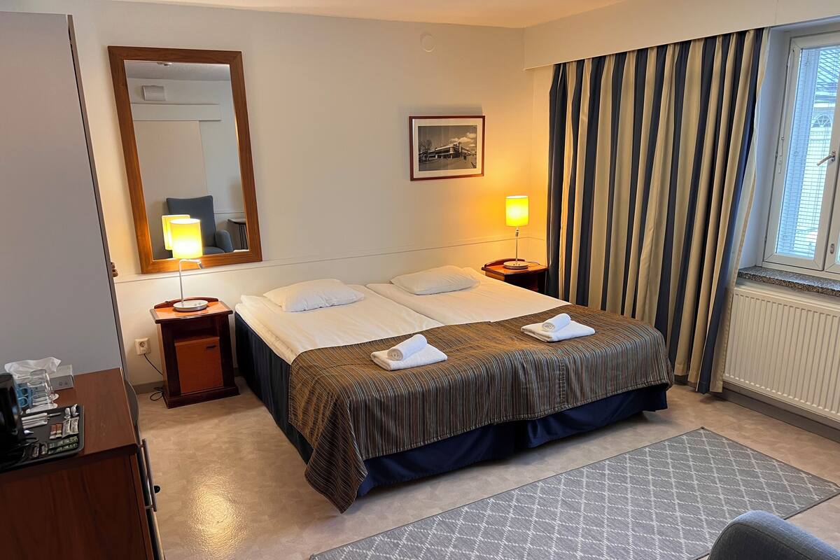 A hotel room with a bed, two bedside lamps, a mirror, a framed picture, and a window with curtains.