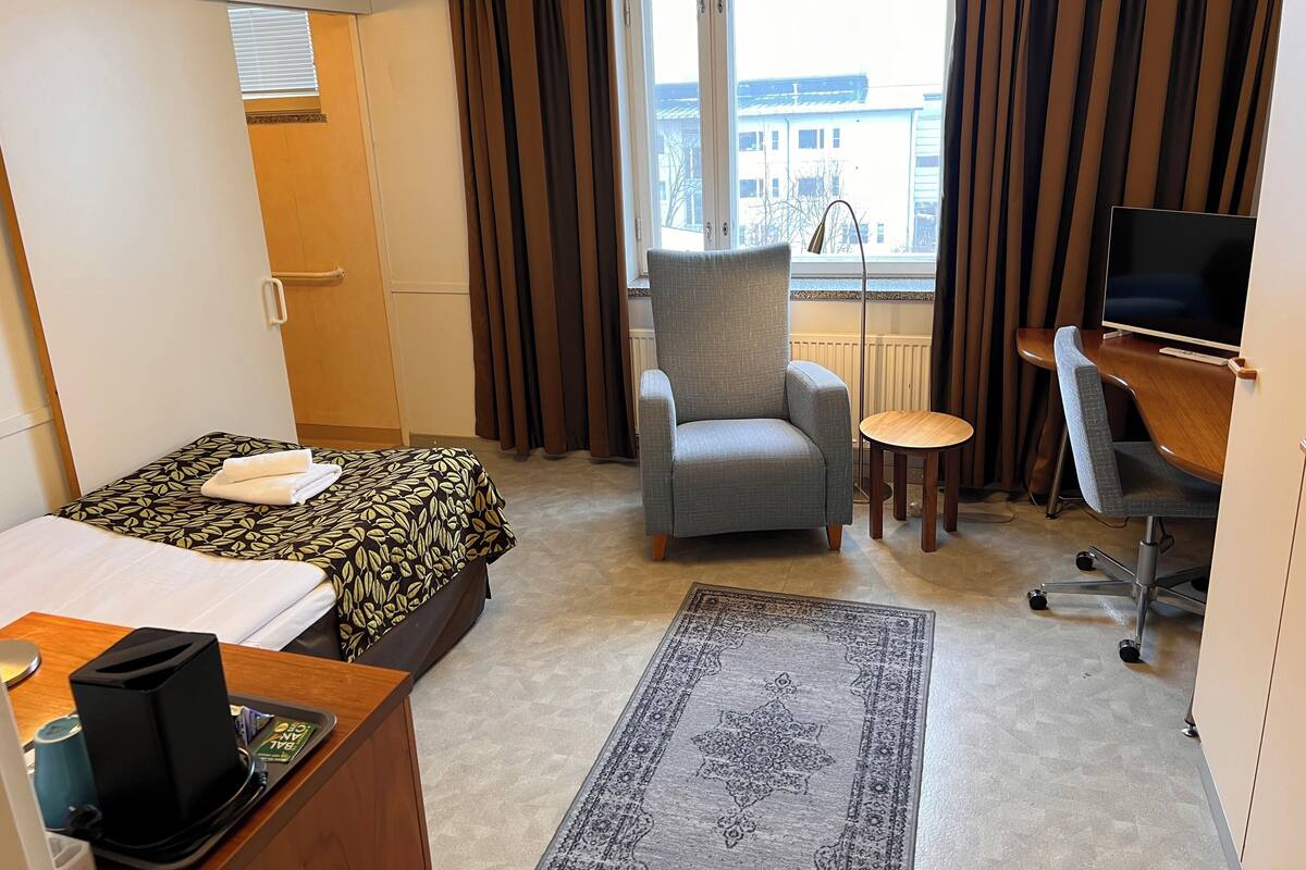 A hotel room with a bed, a grey armchair, a small round table, a desk with a computer, and a large window with curtains.