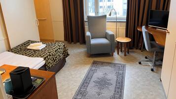 A hotel room with a bed, a grey armchair, a small round table, a desk with a computer, and a large window with curtains.