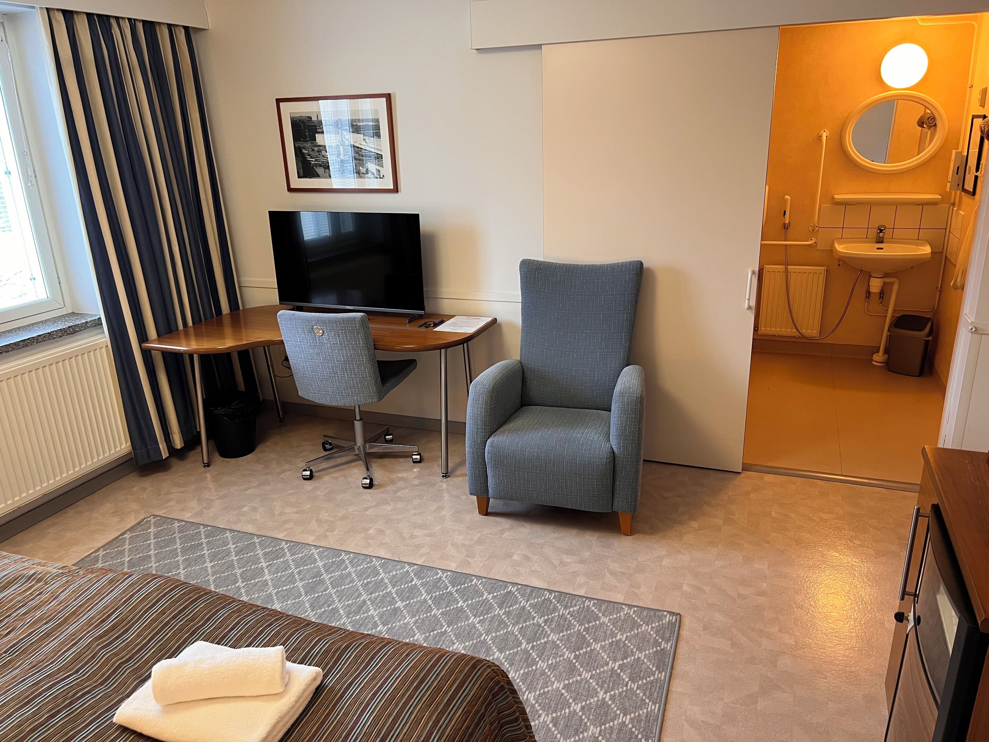 Une chambre d’hôtel comprenant un lit, un bureau avec une chaise, une télévision, une salle de bain avec un lavabo et un miroir, et une fenêtre avec des rideaux.