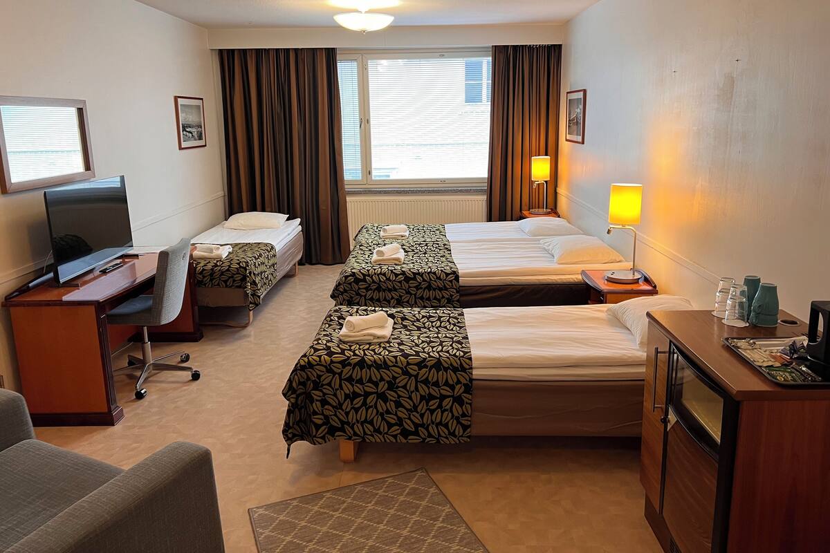 A hotel room with two single beds, a desk with a TV, a chair, a desk lamp, and a small table with glasses and a vase.