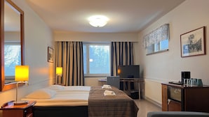 A hotel room with a bed, bedside table, lamp, TV, desk, chair, and a large mirror.