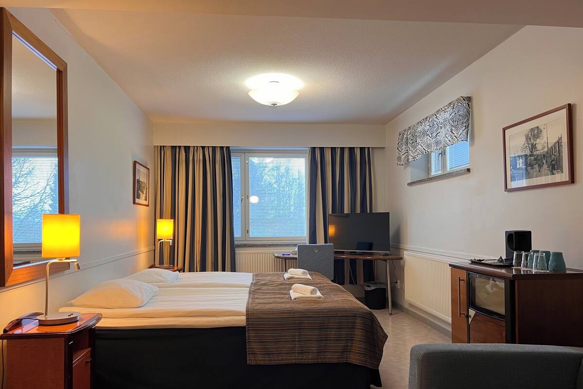 A hotel room with a bed, bedside table, lamp, TV, desk, chair, and a large mirror.