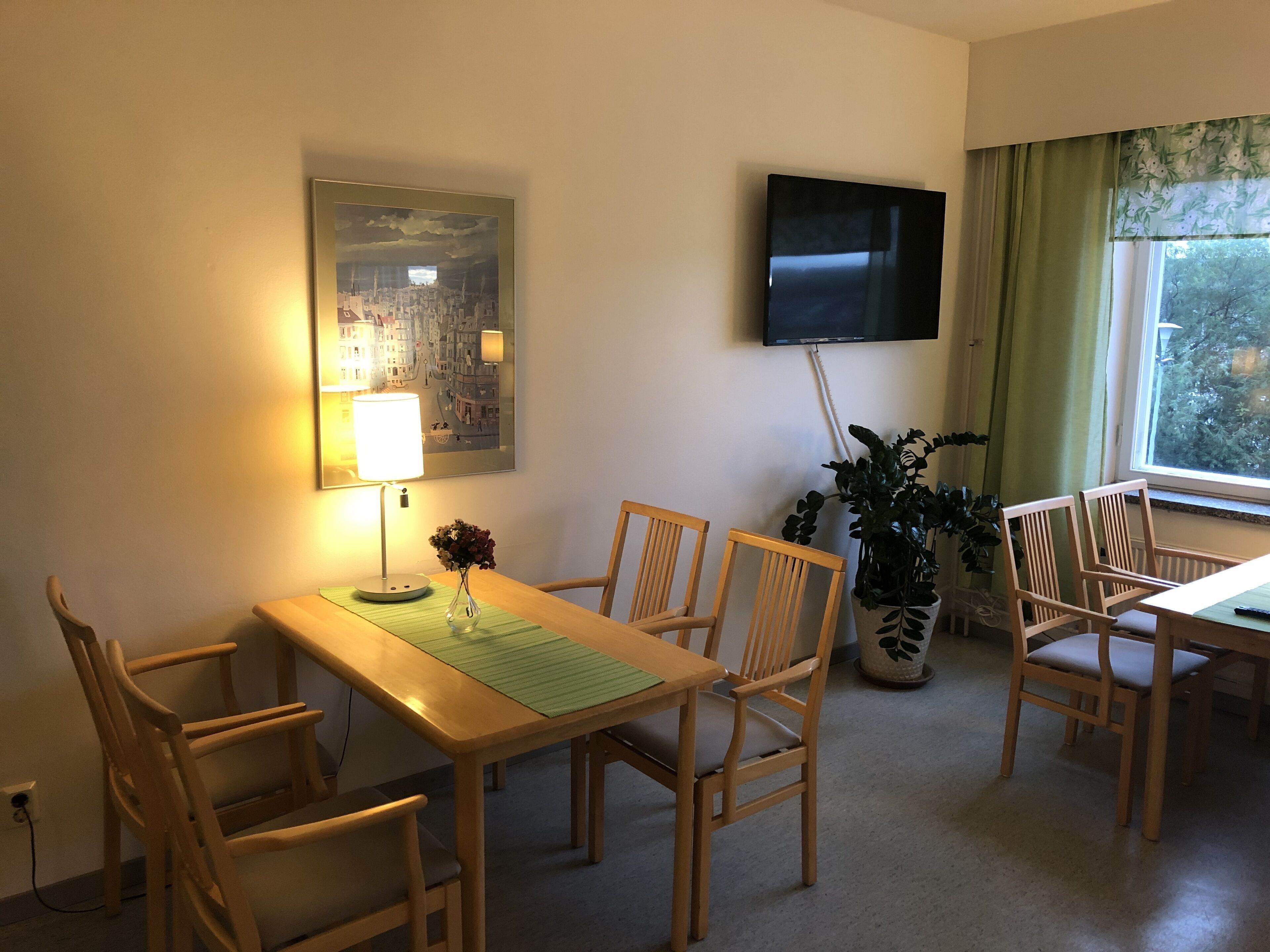 Een eethoek met een tafel, stoelen, een lamp, een televisie en een raam met gordijnen.