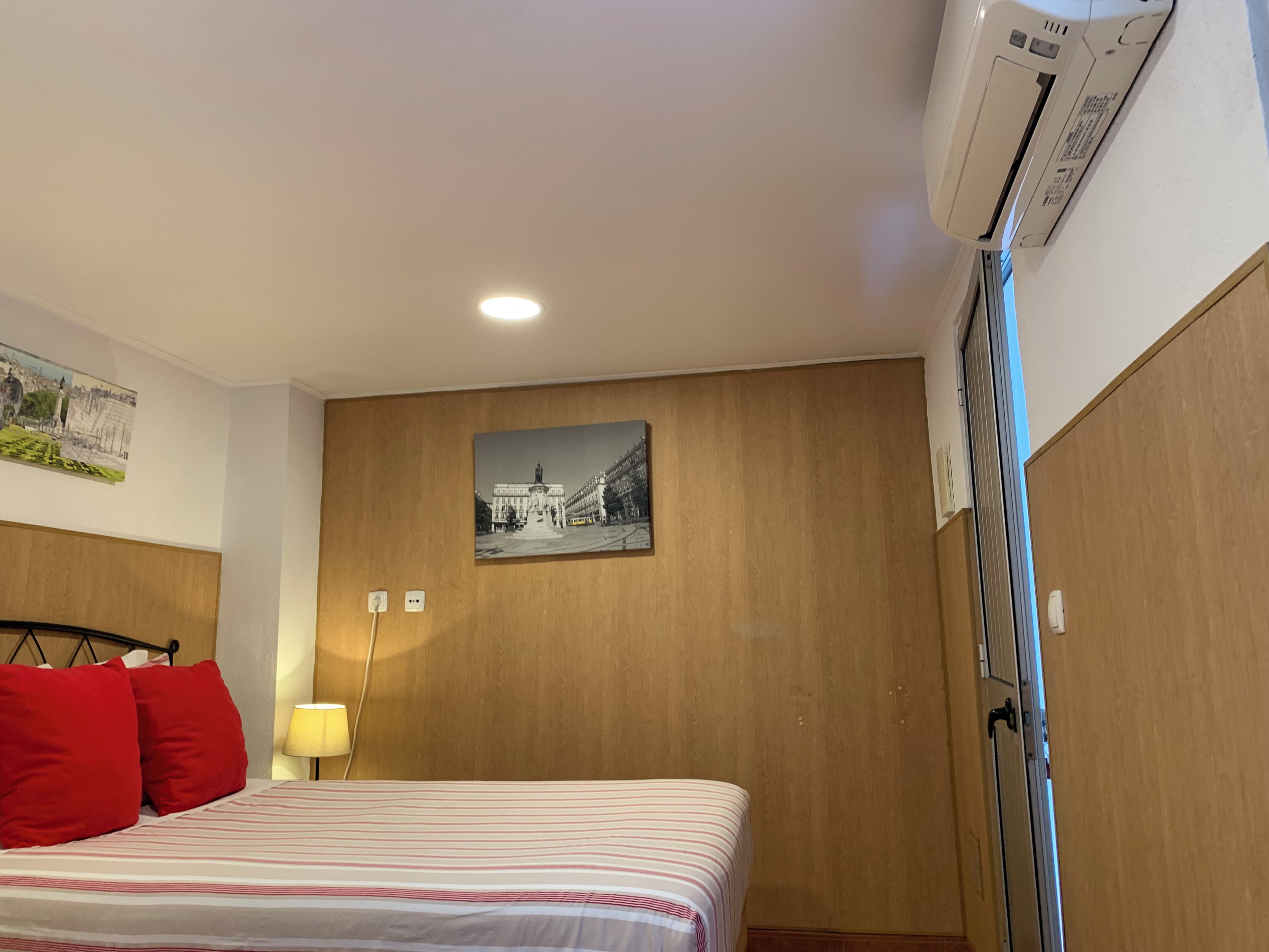 Ein Hotelzimmer mit einem Bett, einer Wandklimaanlage, einer Lampe und einem gerahmten Bild.