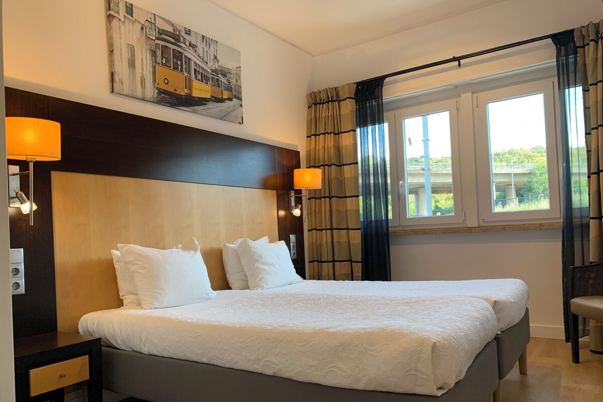 Une chambre d’hôtel avec un grand lit, deux tables de chevet, une tête de lit, un tableau au mur et une fenêtre avec des rideaux.