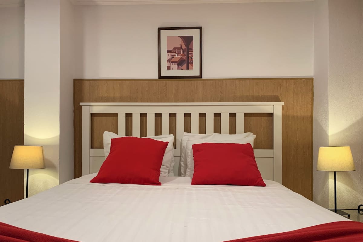 Una cama bien hecha con ropa blanca y dos almohadas rojas, un cabecero de madera y un cuadro enmarcado en la pared.