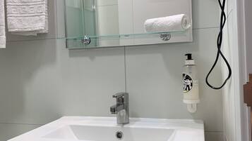 Un baño con un lavamanos blanco, un estante de vidrio, un perchero para toallas, un secador de pelo y un dispensador de jabón.