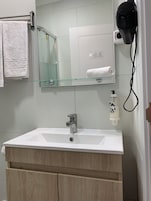Ένα μπάνιο με έναν λευκό νιπτήρα, ένα γυάλινο ράφι, μια κρεμάστρα για πετσέτες, ένα πιστολάκι μαλλιών και ένα δοσομετρητή σαπουνιού.