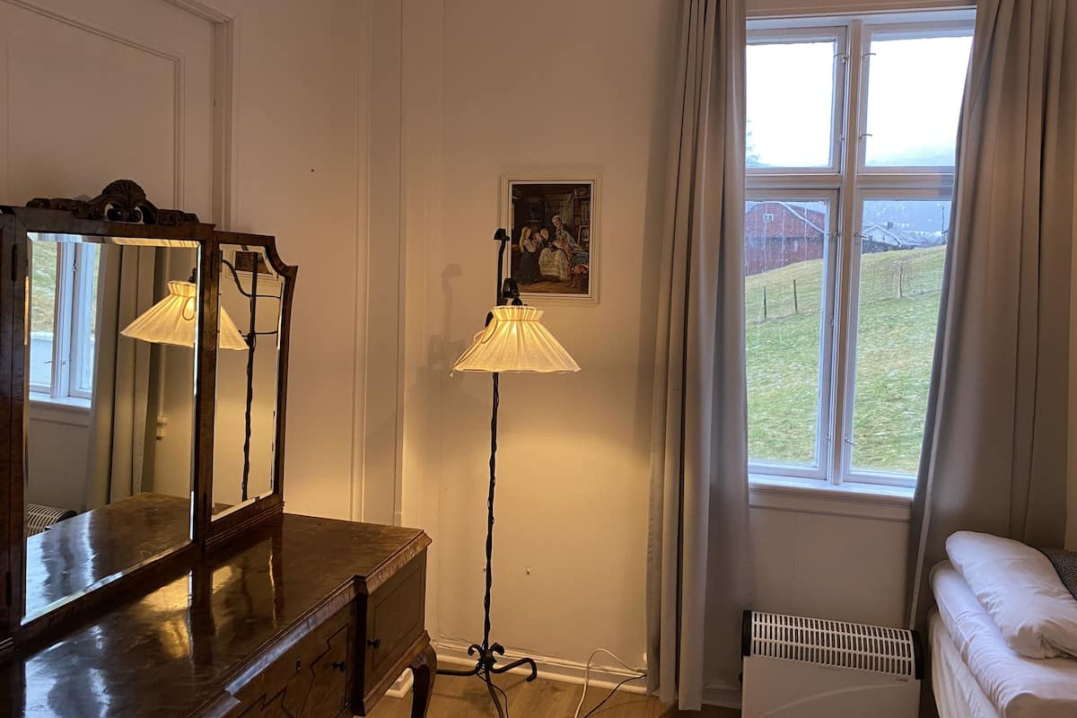Una habitación con un aparador de madera, una lámpara de pie, un radiador, una ventana con cortinas y un cuadro en la pared.