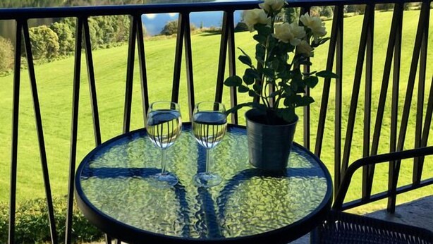 A balcony with a glass table, two chairs, a potted plant, and a view of a lake and mountains. - Lilandtunet Apartments Voss (Voss)