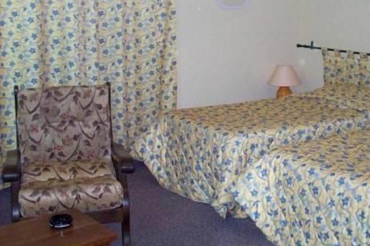 Una habitación de hotel con edredón estampado de flores, una silla, una mesita de noche y una lámpara.