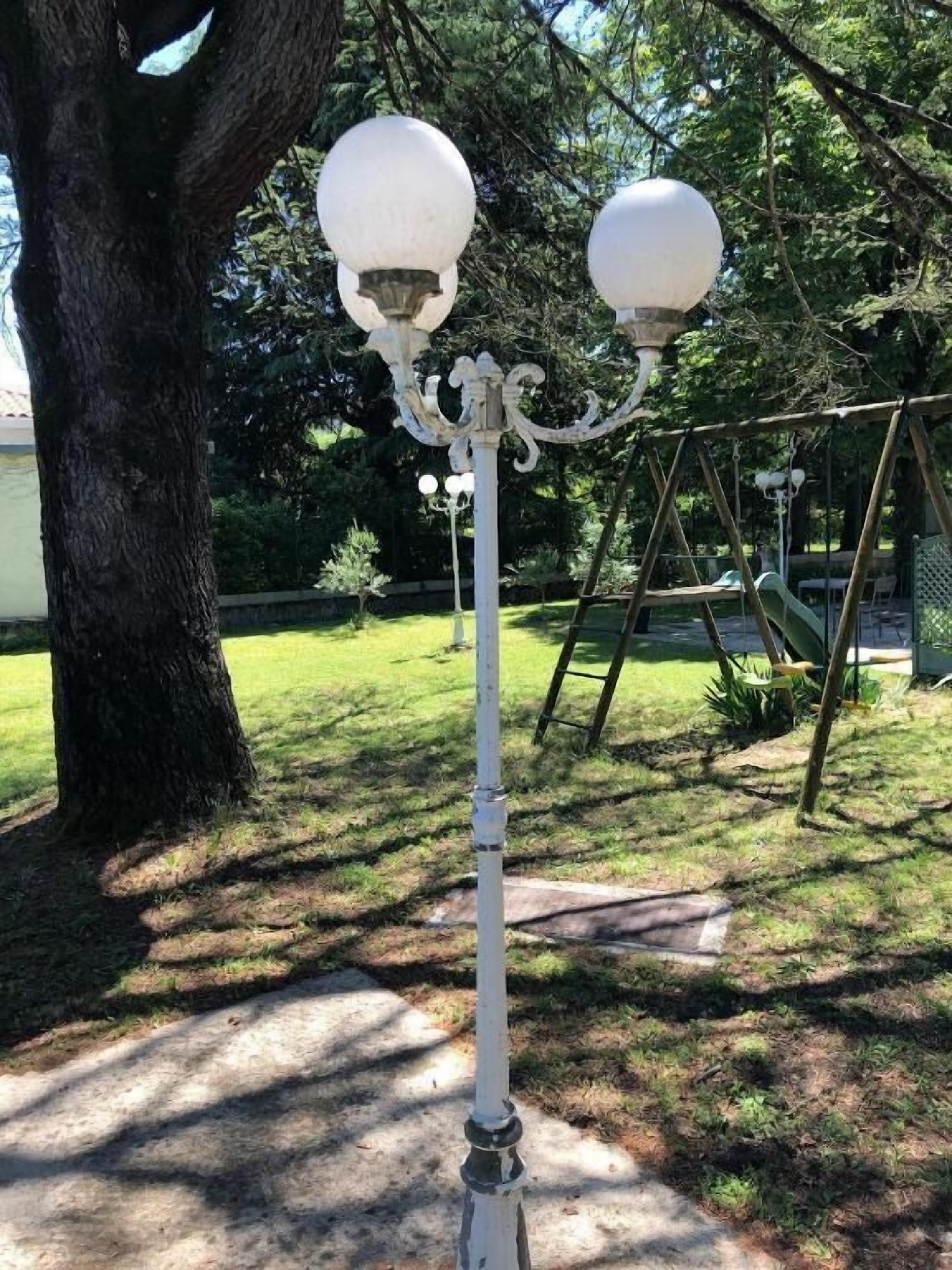 Un poste de luz exterior blanco con dos luces esféricas, ubicado en un área con césped, un columpio y un árbol al fondo.