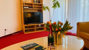 Un salon comprenant une bibliothèque, un téléviseur à écran plat, une table avec un livre et une bouteille, et un vase de tulipes.