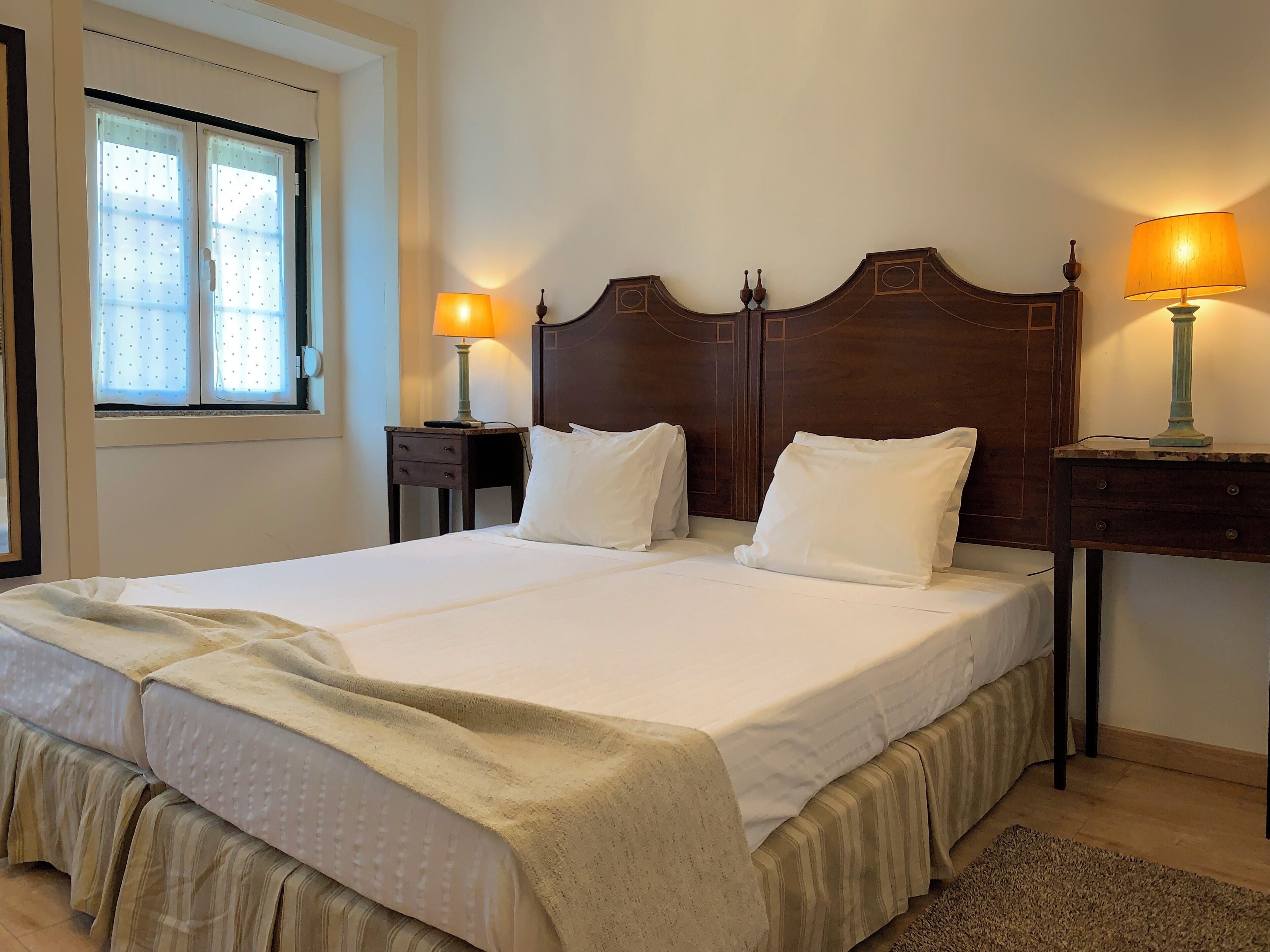 A double bed with a wooden headboard, two bedside tables with lamps, a window with curtains, and a small rug on the floor.