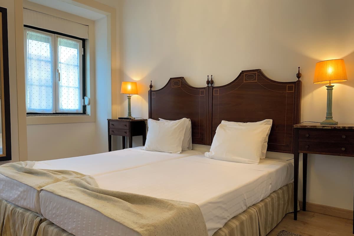 Una cama doble con cabecera de madera, dos mesitas de noche con lámparas, una ventana con cortinas y una pequeña alfombra en el suelo.