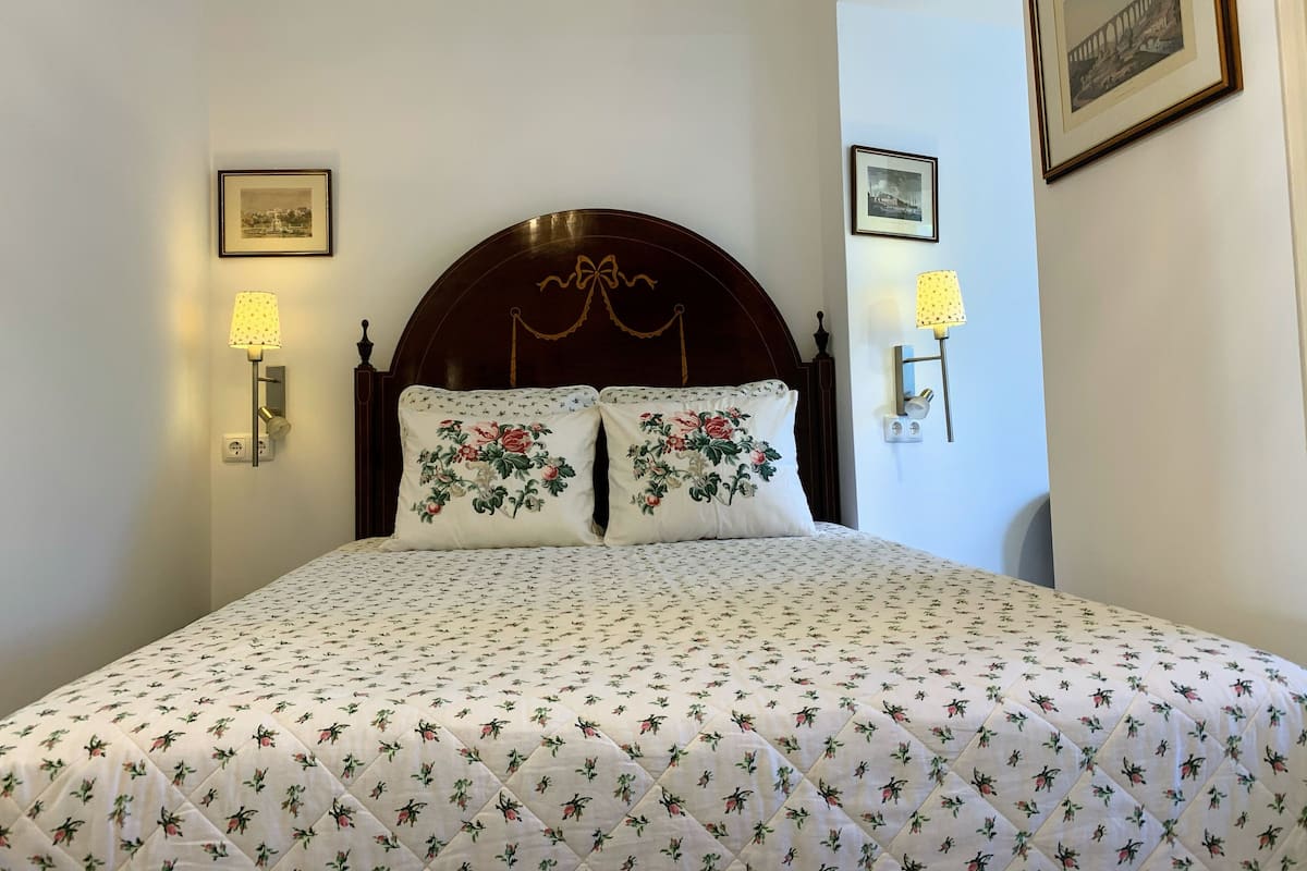 Una cama bien tendida con ropa de cama floral, cabecera de madera con un lazo y lámparas fijadas a la pared.