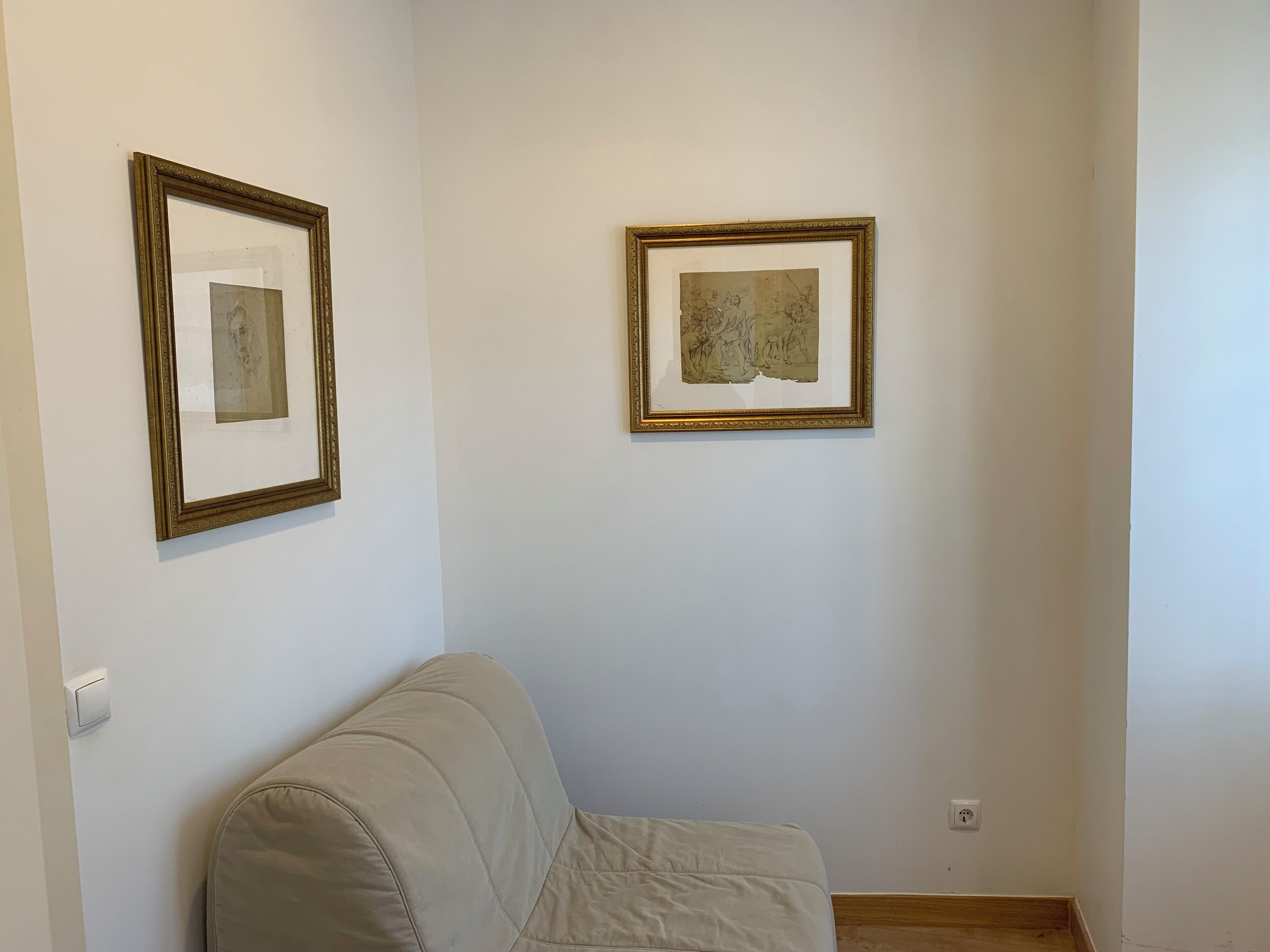 Une pièce avec un canapé beige, deux œuvres d’art encadrées accrochées au mur et un interrupteur.