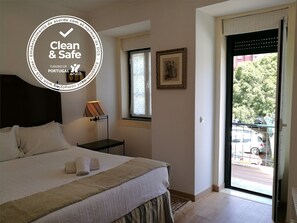 Una habitación de hotel con cama, mesita de noche, lámpara, ventana y puerta que da a un balcón con vistas a coches aparcados y vegetación.