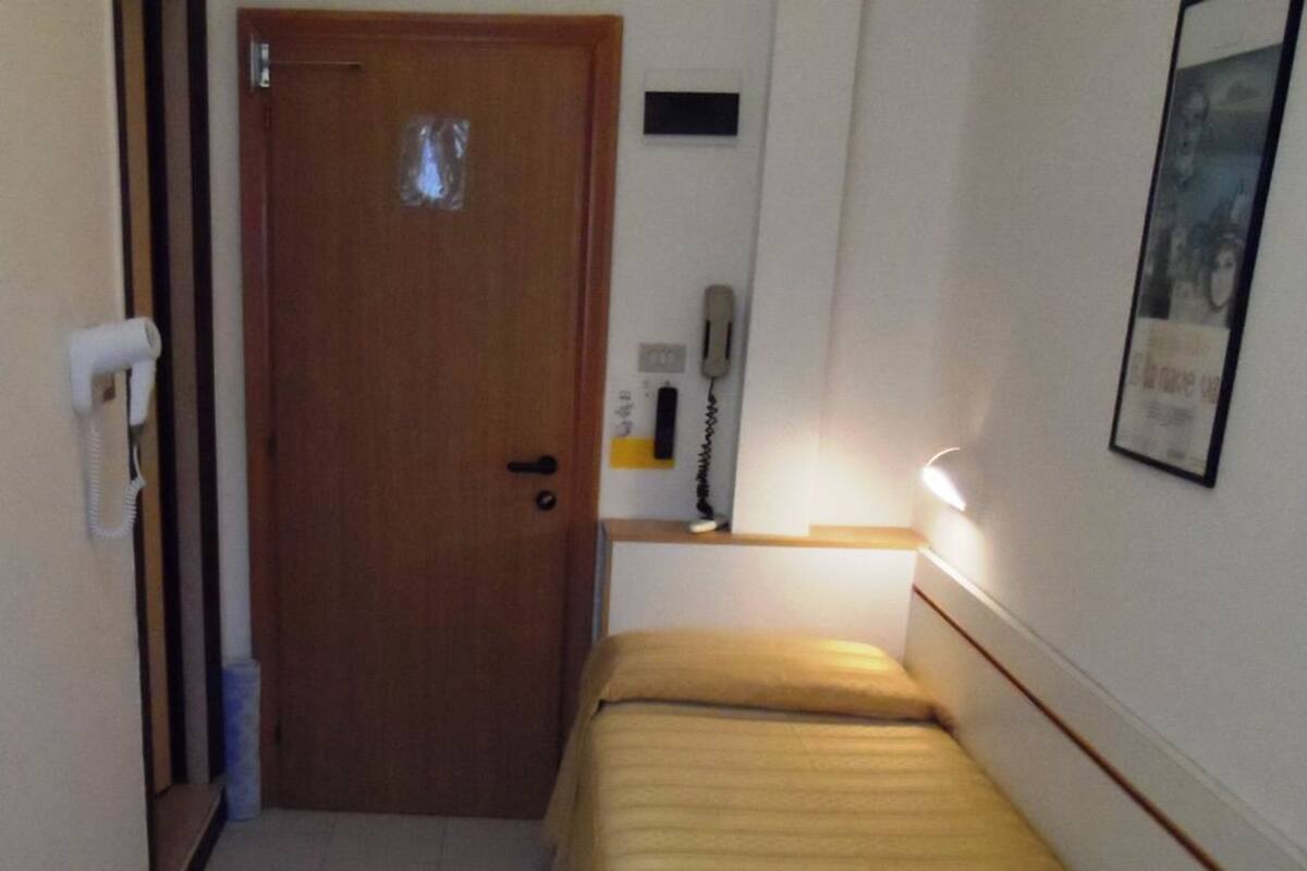 Ein schmaler Flur mit einer Holztür, einem kleinen Nachttisch mit Lampe und einem Bett mit gelber Tagesdecke.