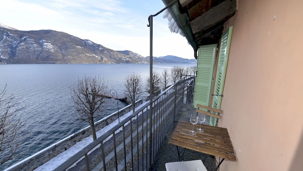 A balcony with a view of a lake and mountains, a wooden table with glasses, and a green shuttered window. - Casa la Finestra sul Lago - WelcHome (Cannobio)