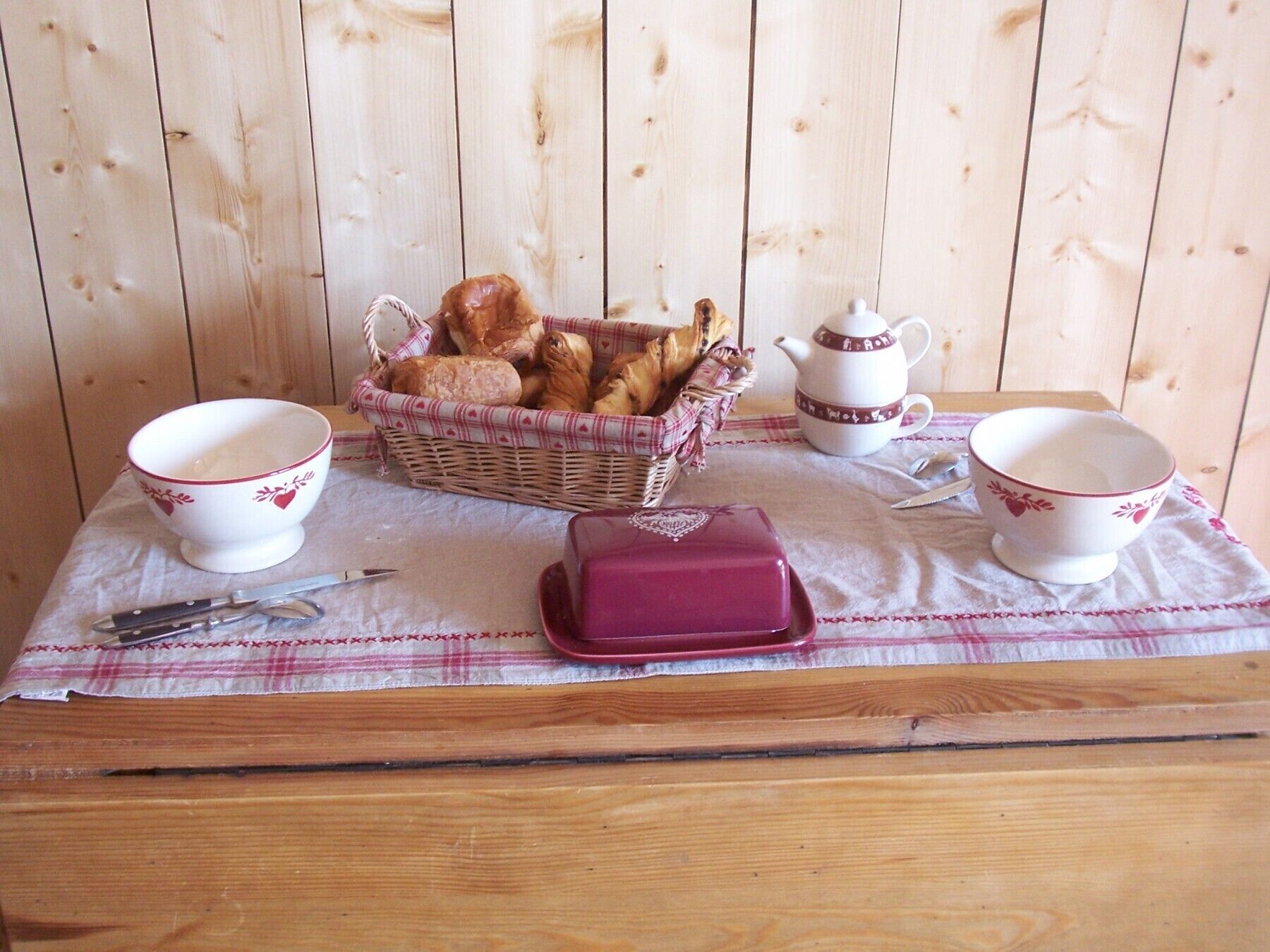 A wooden table with a checkered tablecloth, a wicker basket of bread, two white cups with red patterns, a teapot, and a butter dish.