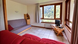 Een kamer met een bed, een slaapbank, een bureau met een computer en een raam met uitzicht op een bos.