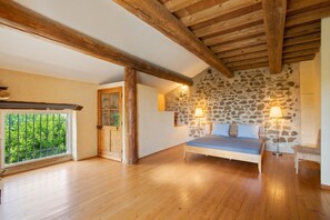 A bedroom with a stone wall, wooden beams, a bed, a chair, and a window with a view of greenery. - Homerez - Villa in Uchaux (Uchaux)