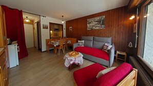 A cozy room with a wooden floor, a sofa, a small table with a red cloth, a dining area with a wooden table and chairs, and a kitchen area with a microwave.