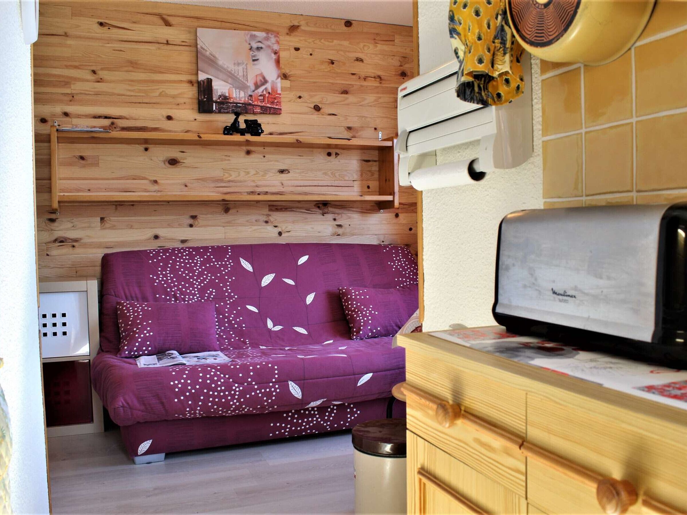 Un espacio habitable pequeño con una pared de madera, un sofá morado con motivos de hojas, un pequeño calentador y un tostador.