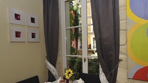 Un coin repas avec une table dressée pour deux personnes, une fenêtre avec des rideaux et une vue sur un bâtiment entouré de verdure.