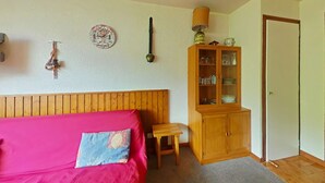 Une chambre à coucher avec une tête de lit en bois, un lit recouvert d’une couverture rouge, une petite table en bois, une armoire aux portes en verre et une porte.
