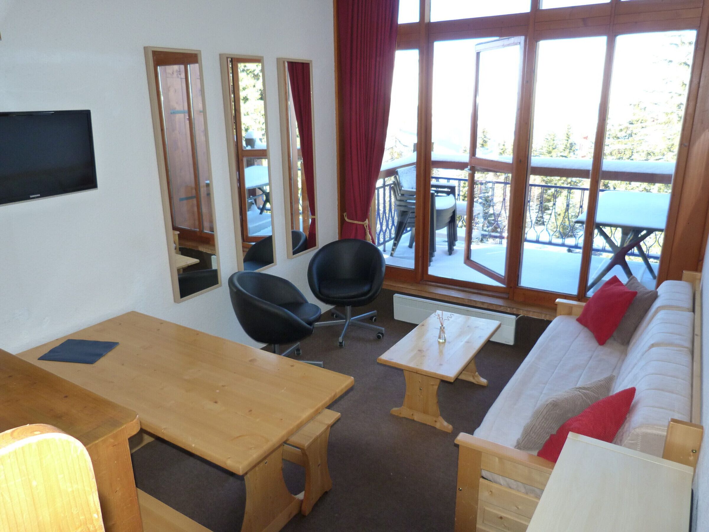 A room with a wooden table, two black chairs, a white sofa with red and grey pillows, a small wooden table, and a TV mounted on the wall.