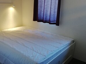 A small, single-bed room with a plain white mattress and a simple, unadorned headboard.