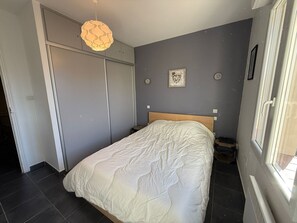 A bedroom with a bed, a wardrobe, a bedside table, a wall-mounted light fixture, and a window.
