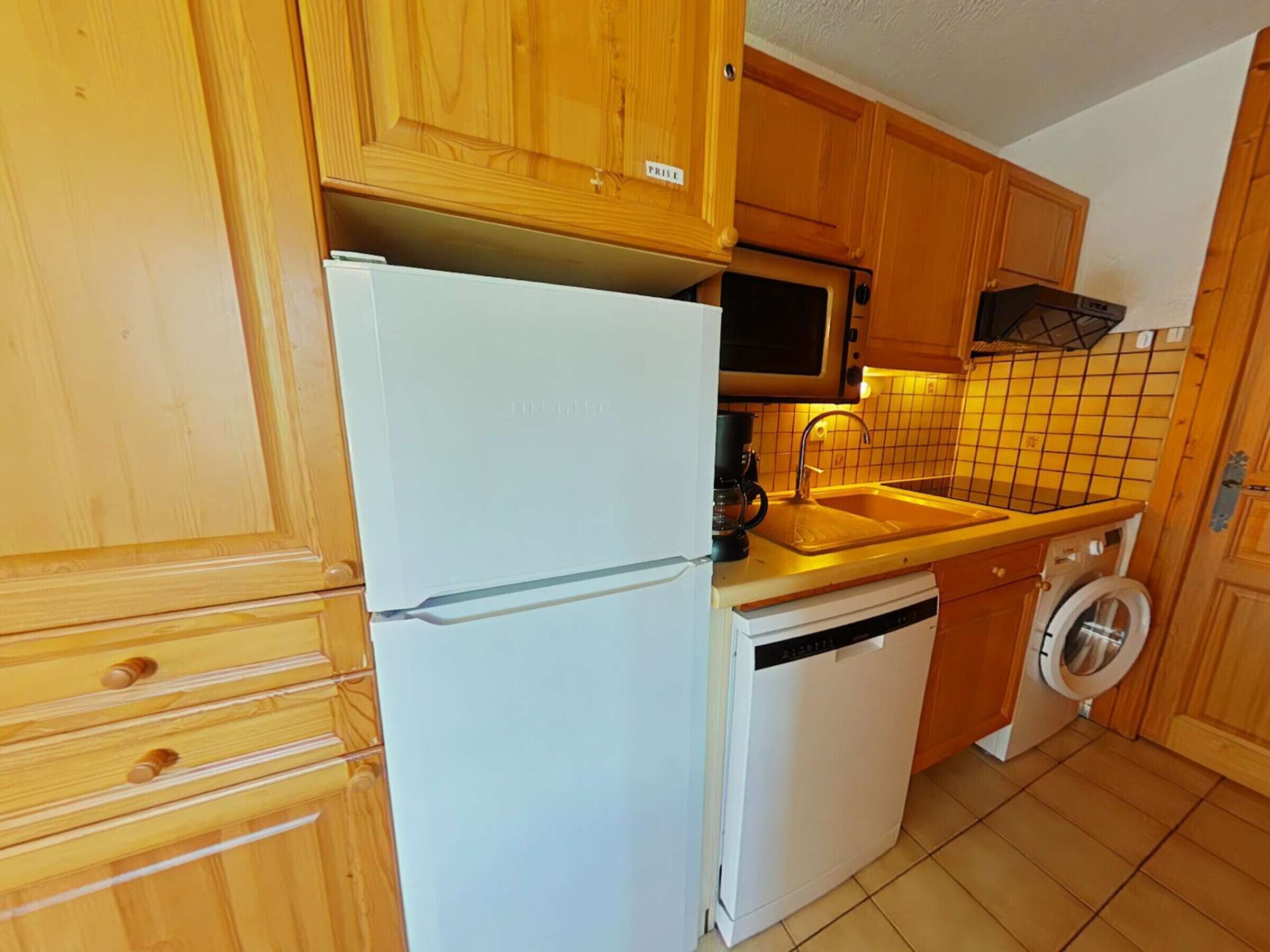Una cocina compacta con armarios de madera, un refrigerador blanco, un microondas, un lavamanos y una lavadora.