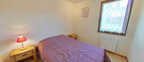 Une chambre à coucher avec un lit, une table de chevet, une lampe, une fenêtre avec des rideaux en dentelle et un radiateur.