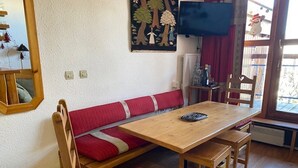 A cozy room with a wooden table, red upholstered bench, and matching chairs. A television is mounted on the wall, and a window provides natural light.
