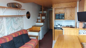 Ett litet rum med en soffa, ett träbord, en köksdel med skåp och en tv, samt en trappa som leder till ett annat rum.