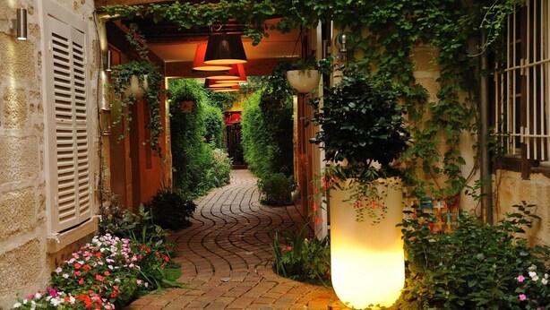 Un étroit sentier pavé de briques, bordé d’une végétation luxuriante et de plantes en fleurs, mène à l’entrée d’un bâtiment, où se trouve une grande colonne illuminée.