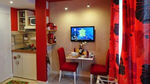 Une cuisine compacte avec un téléviseur fixé au mur, un coin repas avec deux chaises rouges et un rideau rouge orné d’un motif floral.