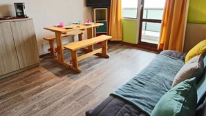 A room with a wooden desk, bench, and a bed with a green blanket. There is a television on the wall and a window with curtains.