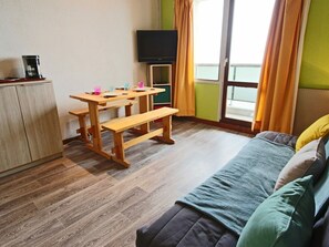 Una habitación con escritorio de madera, banco y cama con manta verde. Hay un televisor en la pared y una ventana con cortinas.