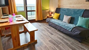 A cozy living room with a wooden floor, a sofa with colorful cushions, a wooden table with plates and glasses, and a television on the left.