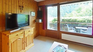 A wooden-paneled room with a television, wooden cabinet, and a sliding glass door leading to an outdoor area with chairs. - Cozy studio - 4 people, parking, pets allowed (Le Grand-Bornand)